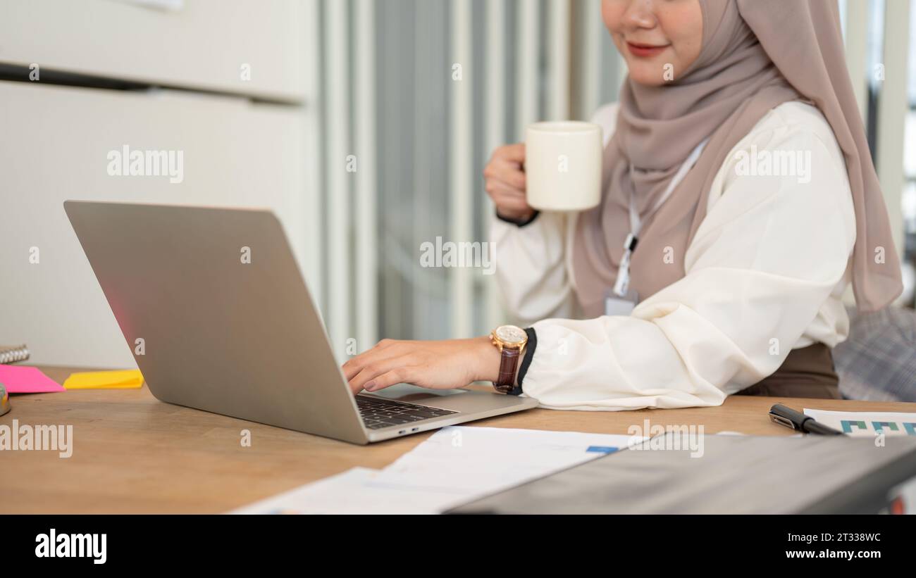 Das zugeschnittene Bild einer attraktiven Millennial-asiatisch-muslimischen Geschäftsfrau trinkt ihren Morgenkaffee, während sie an ihrem Schreibtisch arbeitet Stockfoto Das zugeschnittene Bild einer attraktiven Millennial-asiatisch-muslimischen Geschäftsfrau trinkt ihren Morgenkaffee, während sie an ihrem Schreibtisch arbeitet Stockfoto