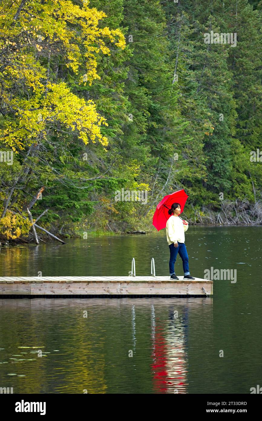 Eine Frau mit einem roten Schirm steht am Ende eines Docks am Robinson Lake in Nord-Idaho. Stockfoto