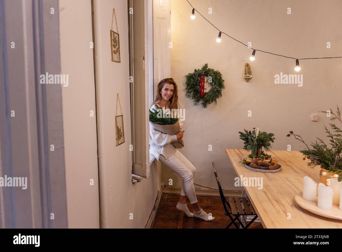 Floristin in weißem Strickpullover, auf Fensterbank sitzend, Weihnachtsstrauß in Handarbeit in Handarbeit in der Werkstatt. Umweltfreundliche Winterdekoration Stockfoto