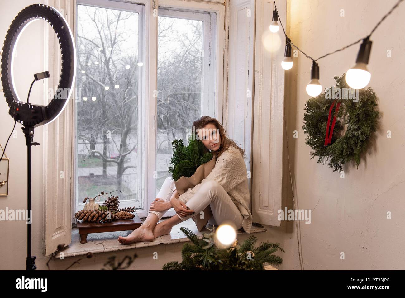 Die junge Bloggerin sitzt auf der Fensterbank in der Nähe des Fensters und hält in der Hand einen Blumenstrauß aus grünen Fichtenzweigen in Handarbeit. Ringlicht-LED-Lampe, Blumenhändler Stockfoto