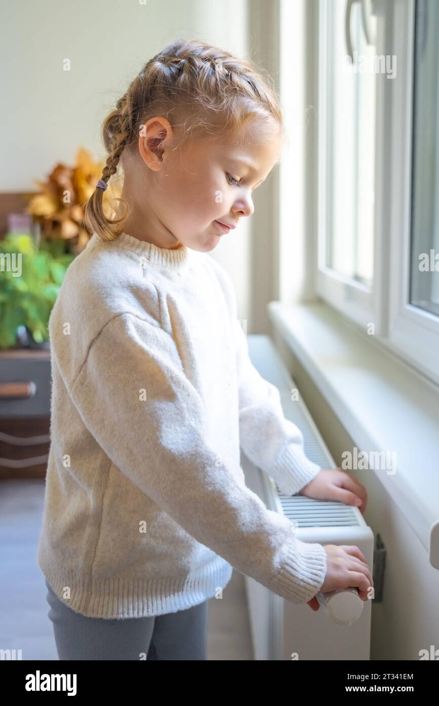 Kleines Mädchen im warmen Pullover, das am Fenster steht, den Batterieheizungsknopf dreht und sich vom Heizkörper aufwärmt. Heizung in einem Apartment Stockfoto