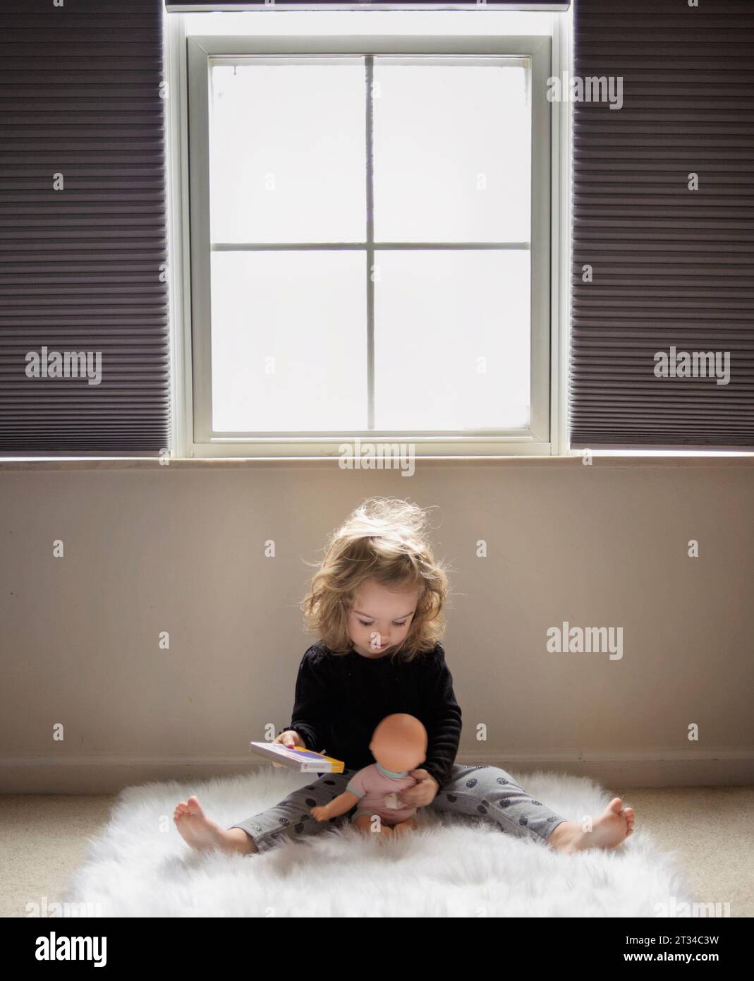 Das kleine Mädchen liest der Babypuppe ein Buch auf einem flauschigen Teppich Stockfoto