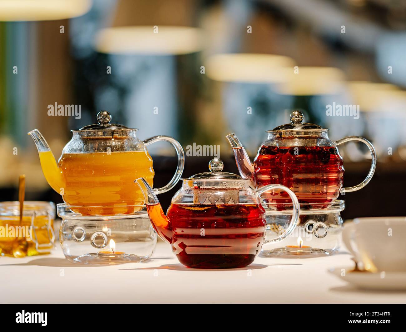 Set aus drei transparenten Glas-Teekannen mit wärmer und Kerzen, heißer Kräutertee auf dem Tisch auf Restaurant-Bokeh-Hintergrund. Mangotee, Hagebutten Kräutertee, würziger Kräutertee mit Sternanis und Honig - Teekonzept Stockfoto