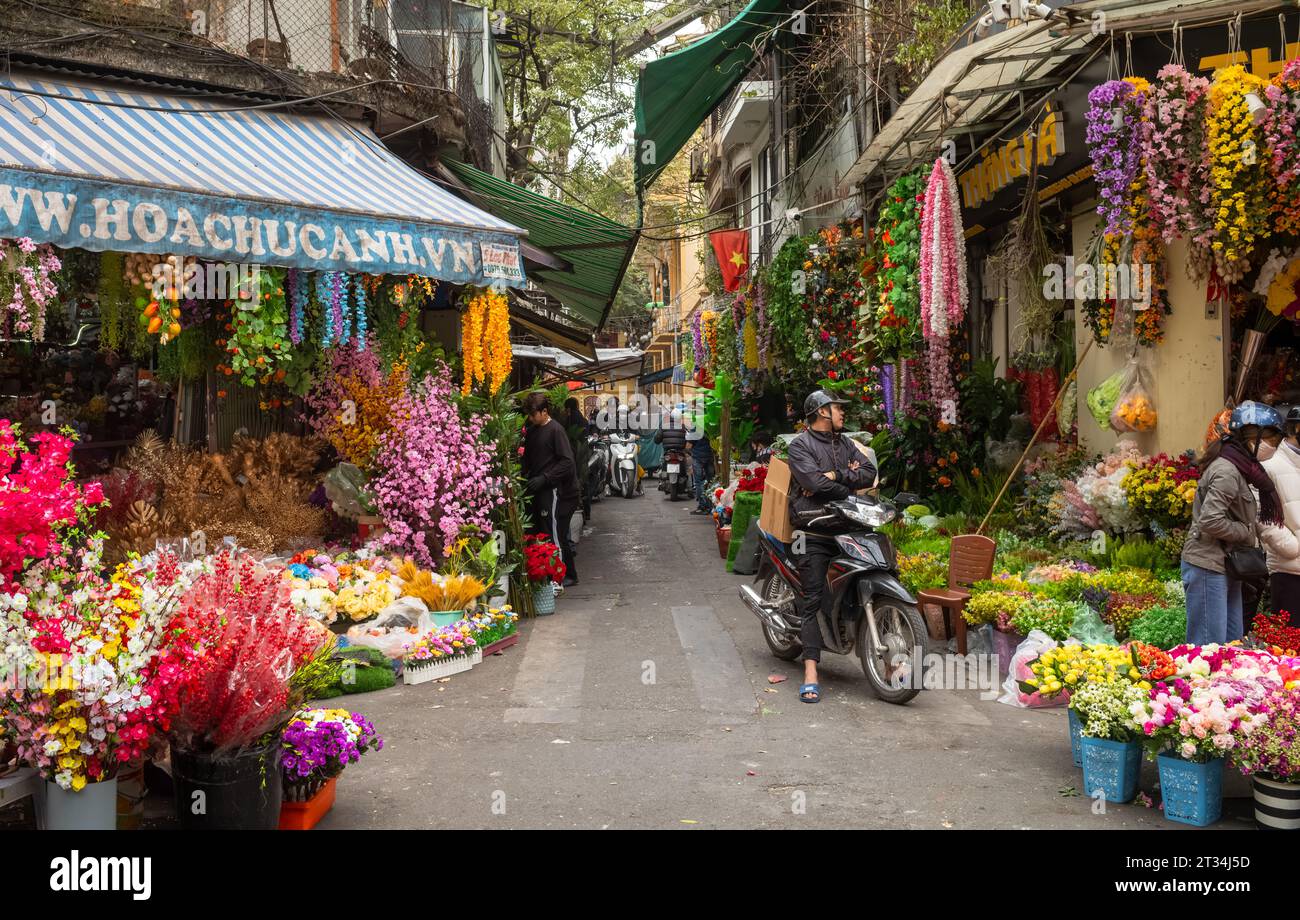 Ein Mann auf einem Motorrad wartet an Geschäften auf beiden Seiten einer Gasse, die künstliche bunte Blumen in der Altstadt von Hanoi, Vietnam, verkauft. Stockfoto
