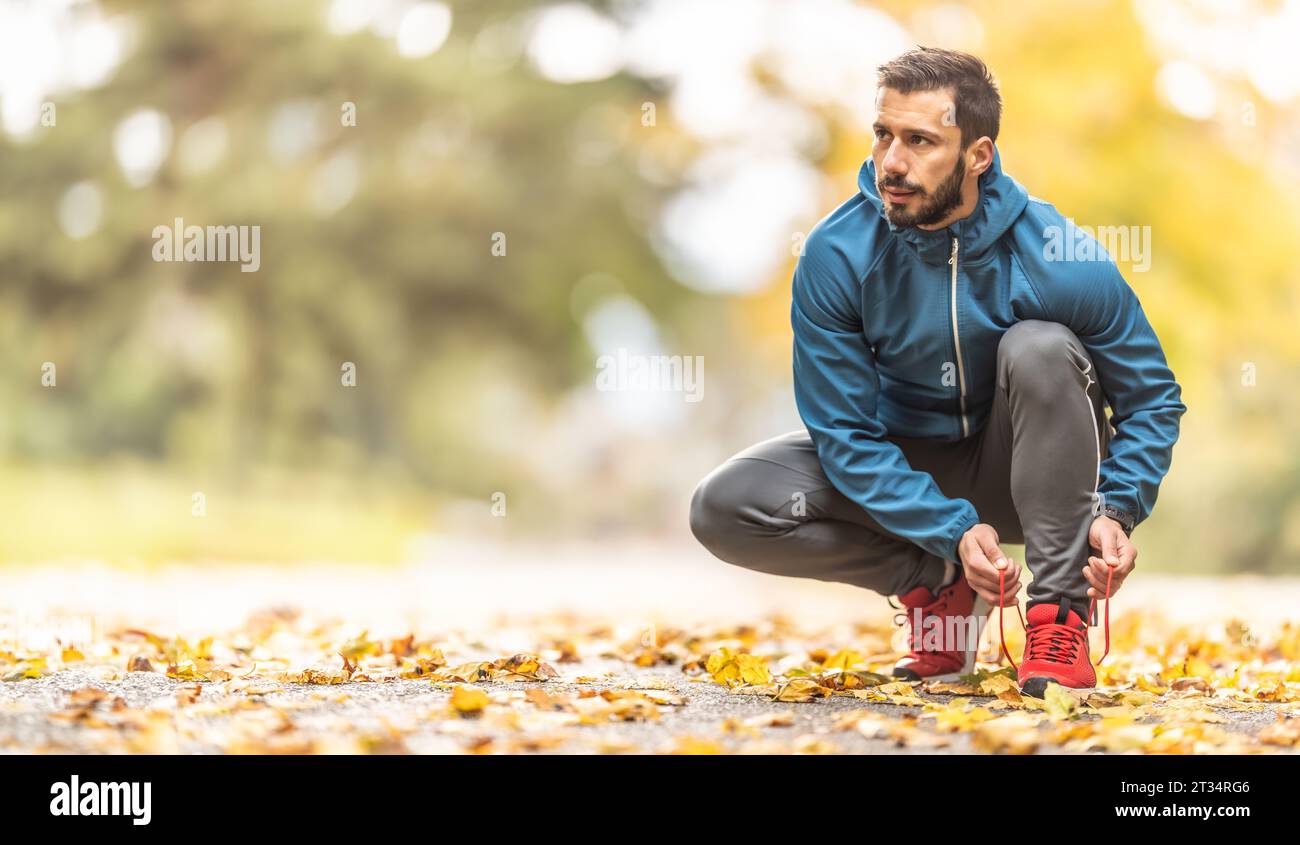 Ein junger Athlet in warmer Sportbekleidung bindet seine Schnürsenkel, während er in einem Herbstpark läuft. Stockfoto