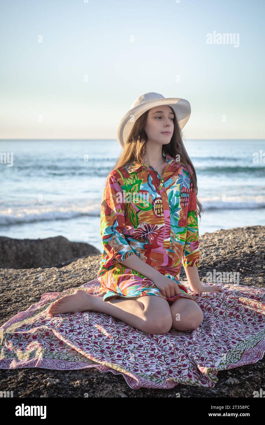 Am späten Nachmittag sitzt eine 20-jährige Kaukasierin auf einem Felsbrocken, die mit einem lebhaften, gemusterten Hemdkleid und einem schicken Hut verziert ist Stockfoto