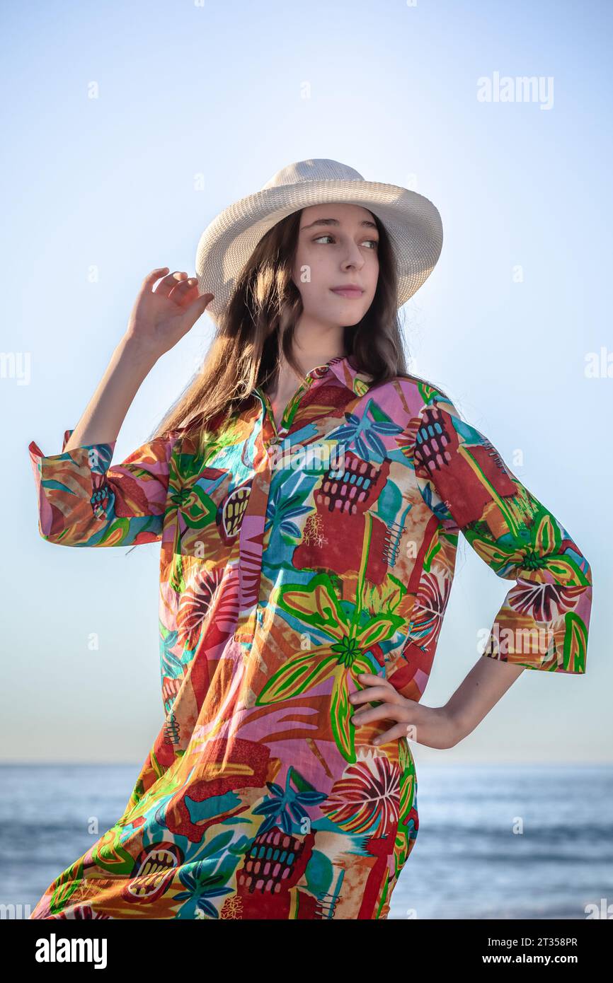 Eine Nahaufnahme fängt den strahlenden Charme einer jungen kaukasischen Frau in einem farbenfrohen Hemdkleid und einem stilvollen Hut am Strand ein Stockfoto