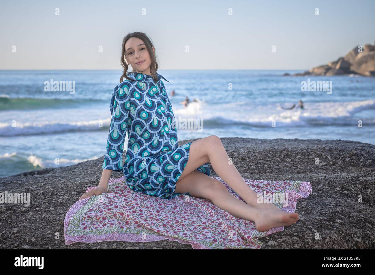 Am späten Nachmittag sitzt eine 20-jährige Kaukasierin auf einem Felsbrocken, die in einem lebhaften, gemusterten Hemdkleid verziert ist Stockfoto