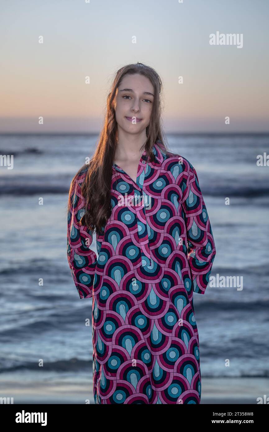 Eine 20-jährige Kaukasierin steht bei Sonnenuntergang am Strand und trägt ein lebendiges und farbenfrohes Hemdkleid, während die Sonne ein warmes Licht über den Strand wirft Stockfoto