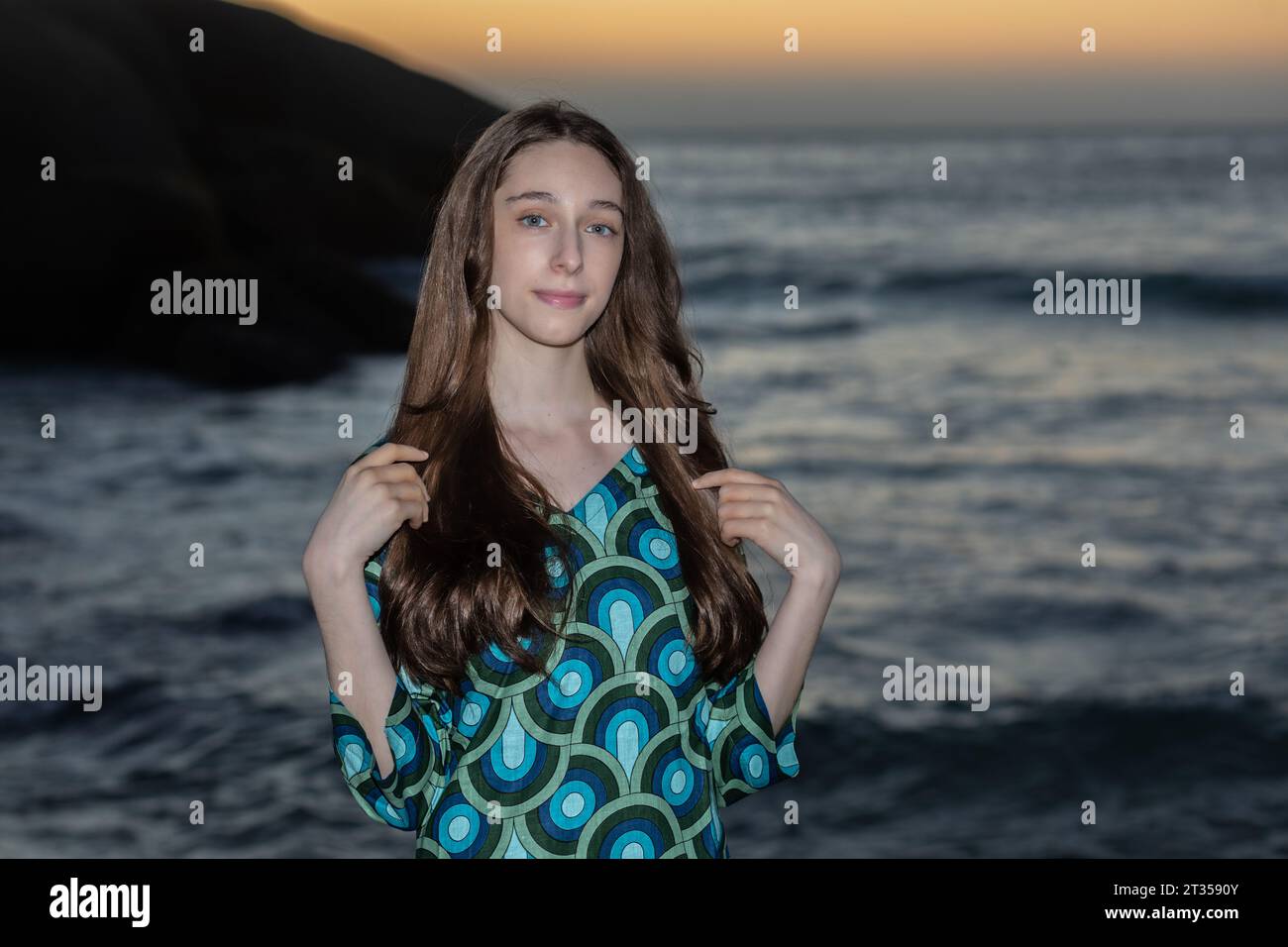 Eine 20-jährige Kaukasierin steht bei Sonnenuntergang am Strand und trägt ein lebendiges und farbenfrohes Hemdkleid, während die Sonne ein warmes Licht über den Strand wirft Stockfoto
