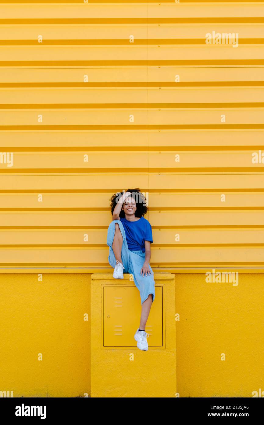 Hübsche Frau, die auf der Plattform vor der gelben Wand sitzt und lächelt Stockfoto