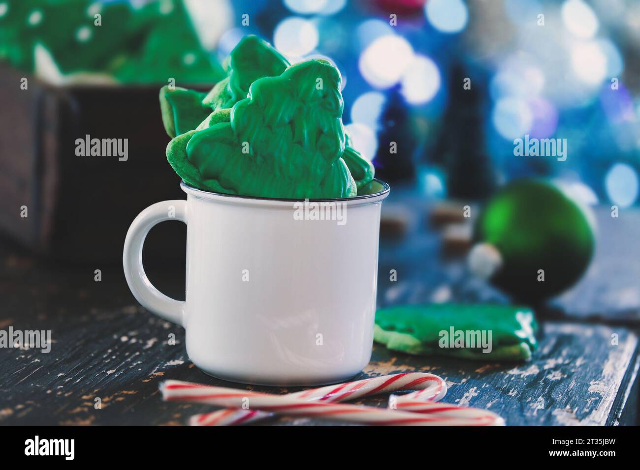 Milchkekse in Form eines Weihnachtsbaums mit grüner Glasur in einer weißen Kaffeetasse mit Zuckerstangen in der Nähe. Selektiver Fokus mit verschwommenem f Stockfoto