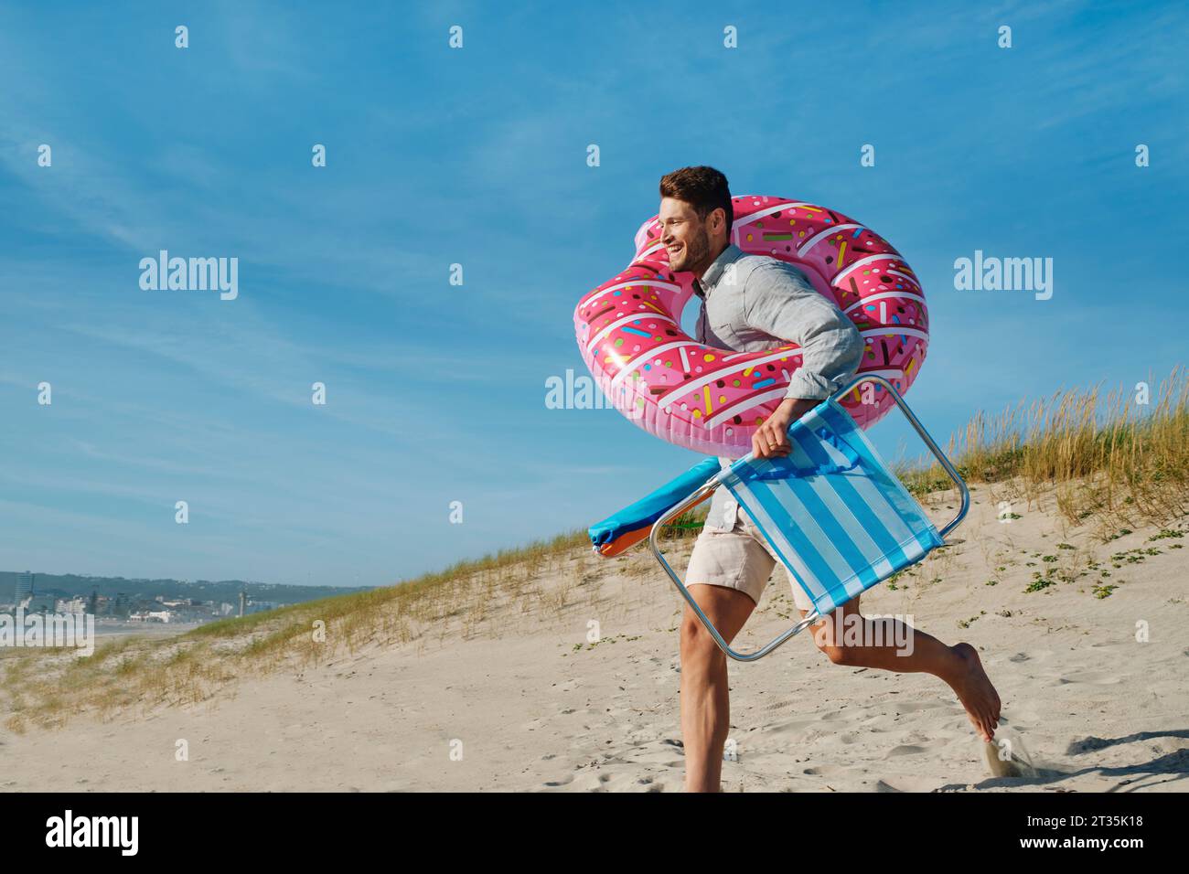 Glücklicher Mann, der am sonnigen Tag mit aufblasbarem Schwimmring und Klappstuhl am Strand läuft Stockfoto