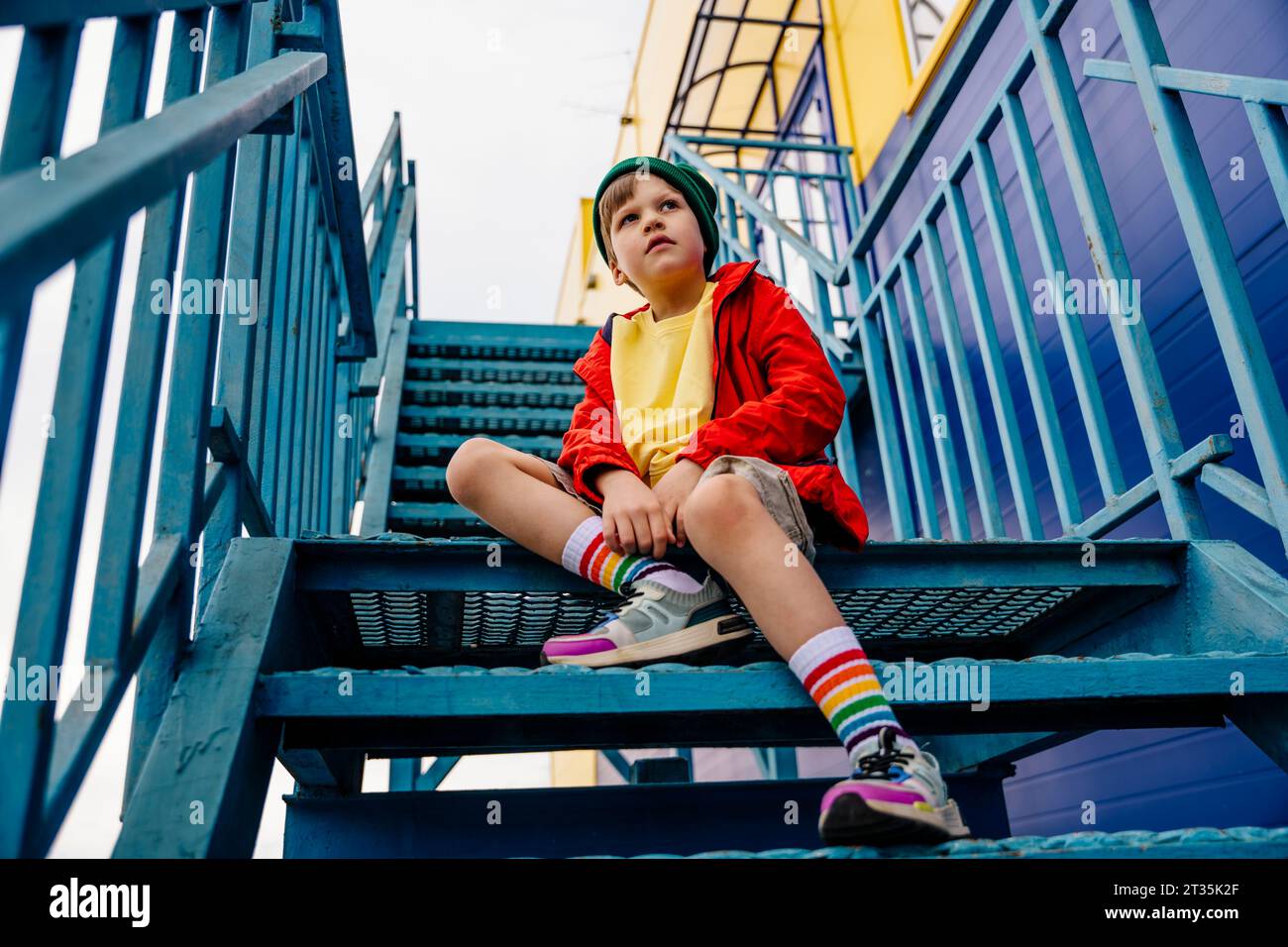 Nachdenklicher Junge in roter Jacke, der auf einer Metalltreppe sitzt Stockfoto