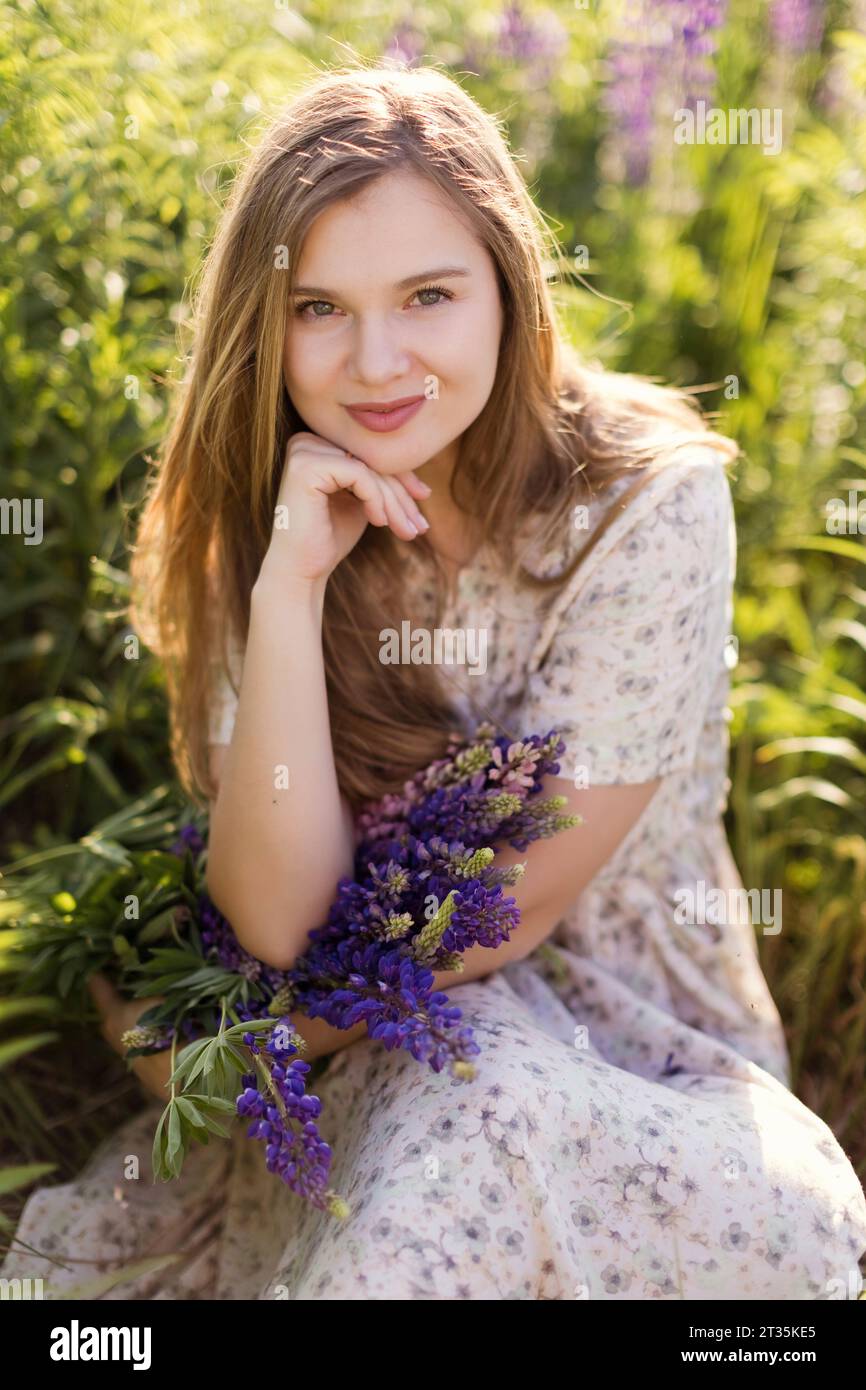 Lächelnde Frau mit Hand am Kinn und lila Lupinenblüten, die auf dem Feld sitzen Stockfoto