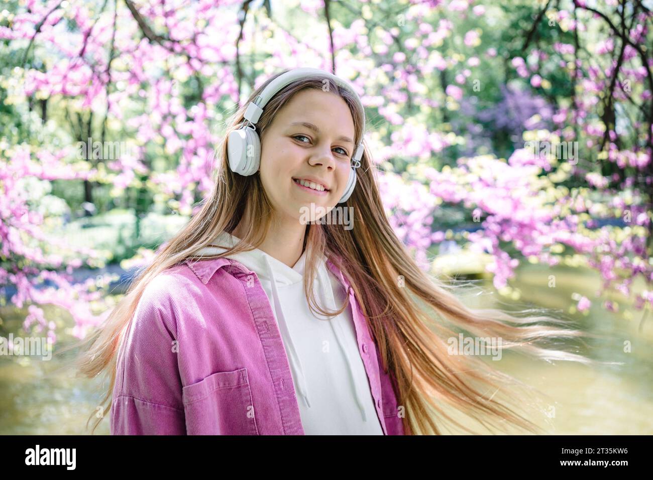 Lächelndes Teenager-Mädchen mit langen blonden Haaren, das Musik mit Kopfhörern am See im Park genießt Stockfoto