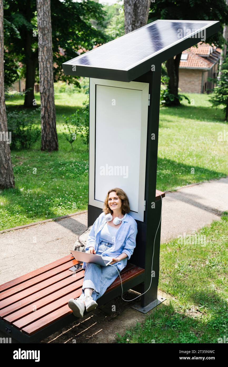 Lächelnde Frau mit Laptop, die auf einer Bank in der Nähe der Solarladestelle im Park sitzt Stockfoto