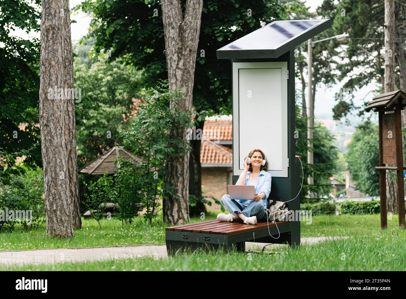 Frau, die auf der Bank sitzt und Musik in der Nähe der Solarladestelle im Park hört Stockfoto