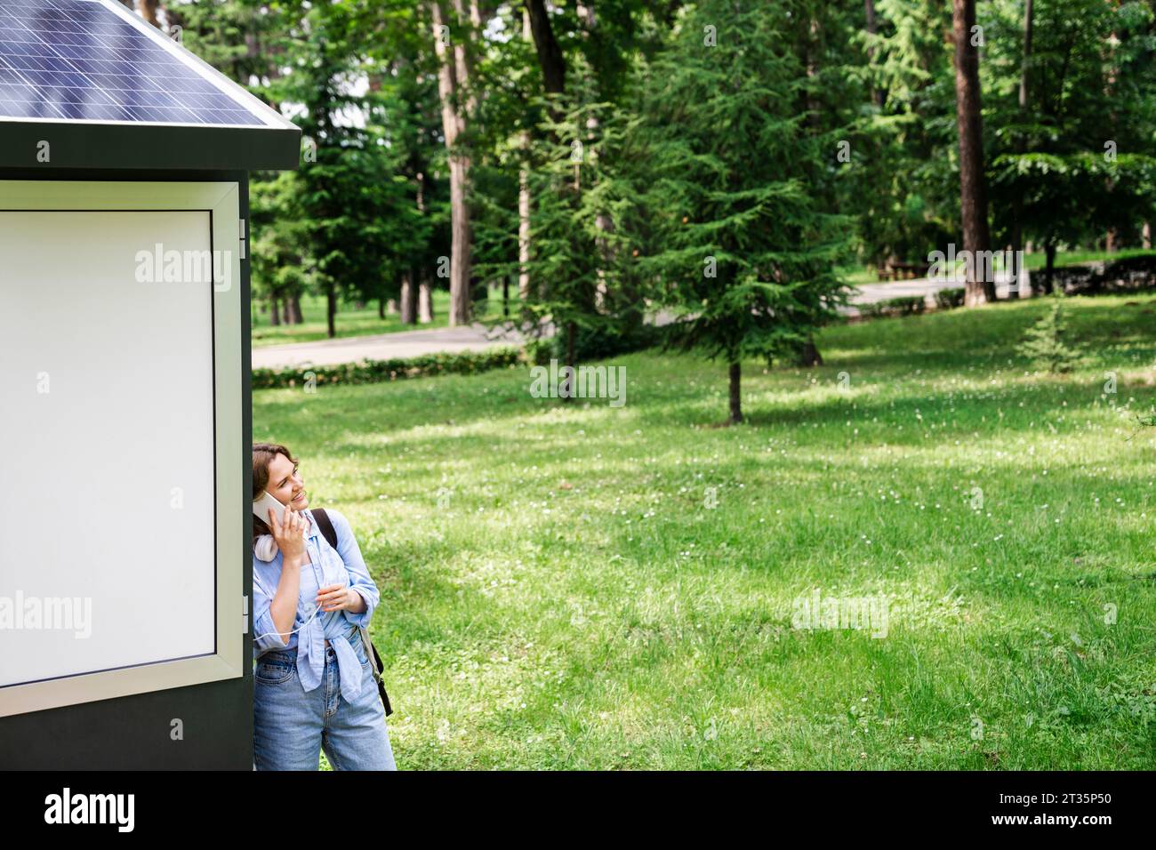 Frau, die auf dem Handy spricht, lehnt sich an die Solarladestelle im Park Stockfoto
