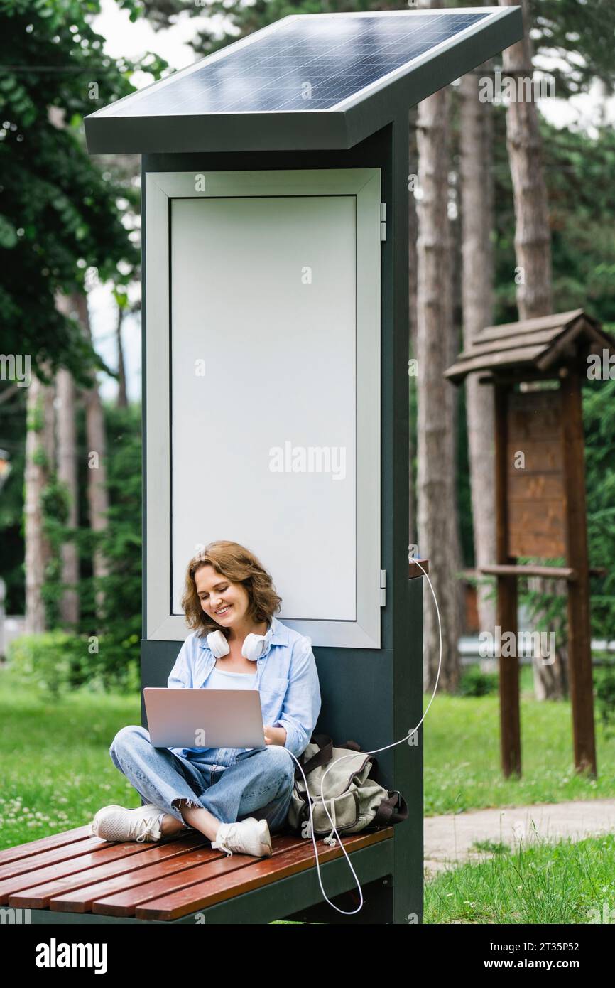 Lächelnde Frau, die einen Laptop benutzt und auf einer Bank in der Nähe des Solarladesteckes im Park sitzt Stockfoto