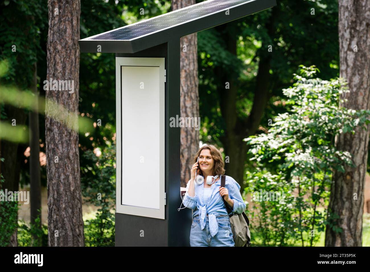 Lächelnde Frau, die auf dem Smartphone steht, steht in der Nähe der Solarladestelle im Park Stockfoto