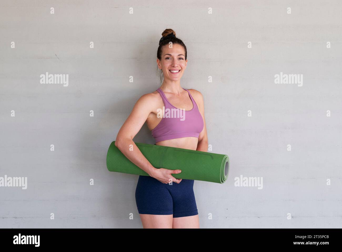 Lächelnde Frau, die eine Yogamatte hält, die vor einer Betonwand steht Stockfoto