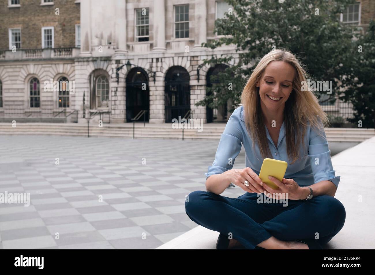 Lächelnde Geschäftsfrau, die ihr Smartphone benutzt und im Kreuz auf der Bank sitzt Stockfoto