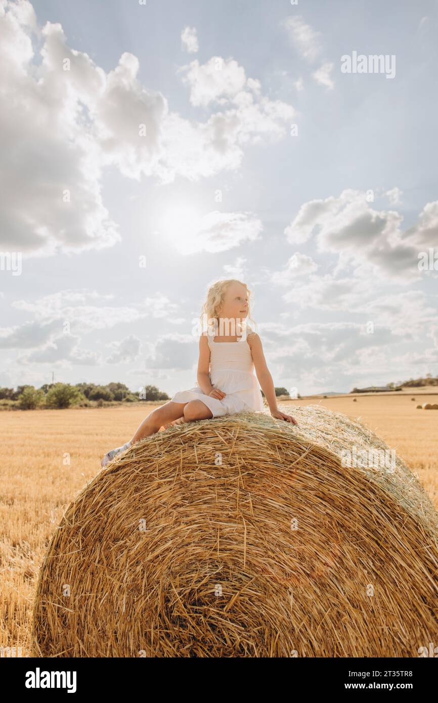 Besinnliches blondes Mädchen, das am sonnigen Tag auf Strohballen sitzt Stockfoto