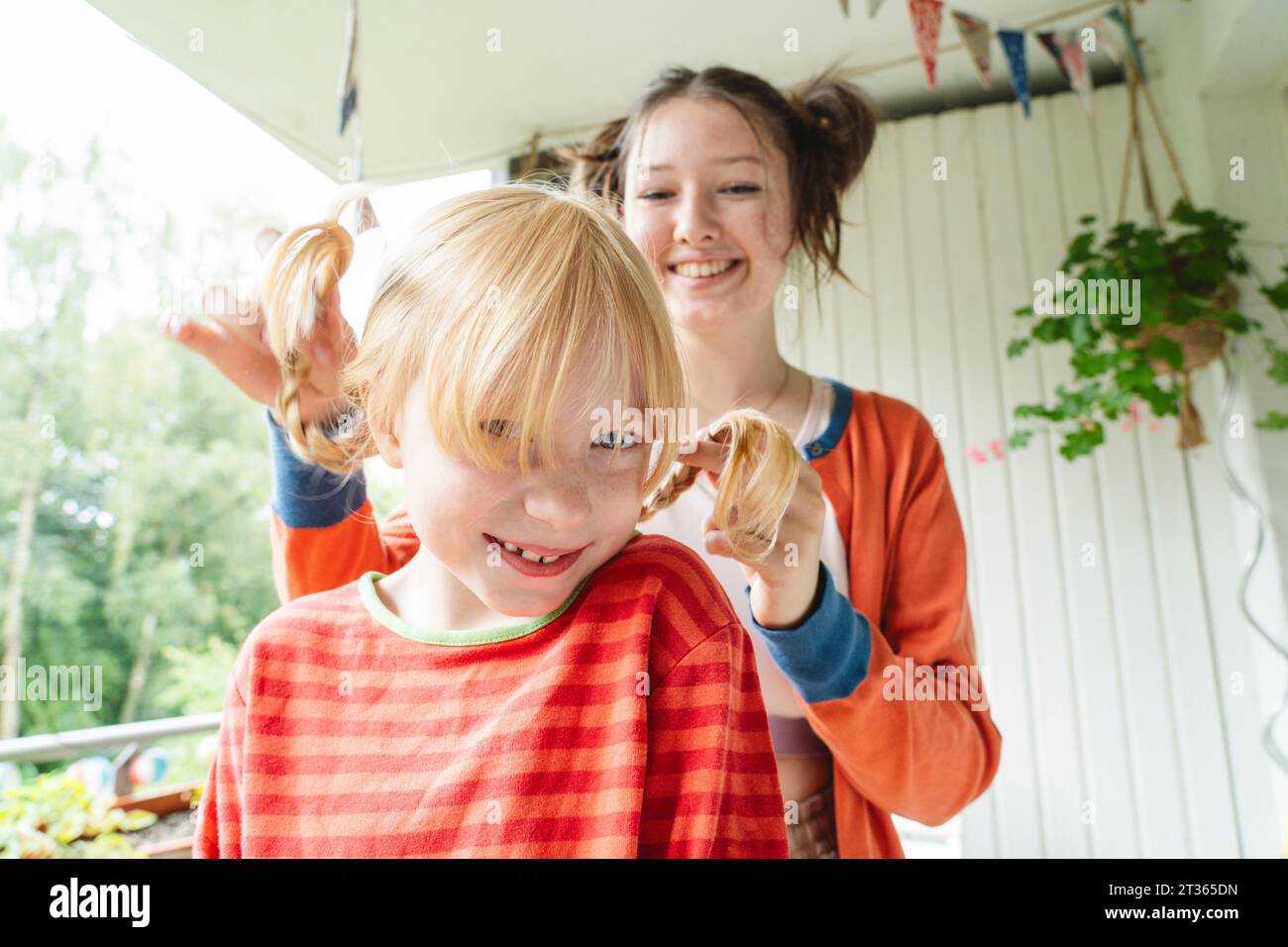 Blondes Mädchen, das von ihrer glücklichen Teenager-Schwester auf dem Balkon geflochten wird Stockfoto