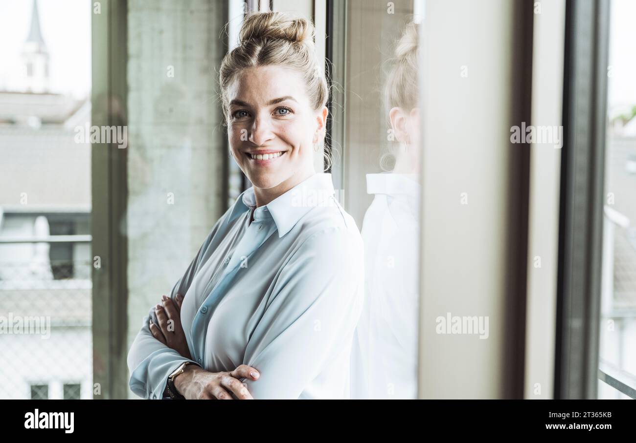 Glückliche Geschäftsfrau, die mit gekreuzten Armen in der Nähe des Fensters im Büro steht Stockfoto