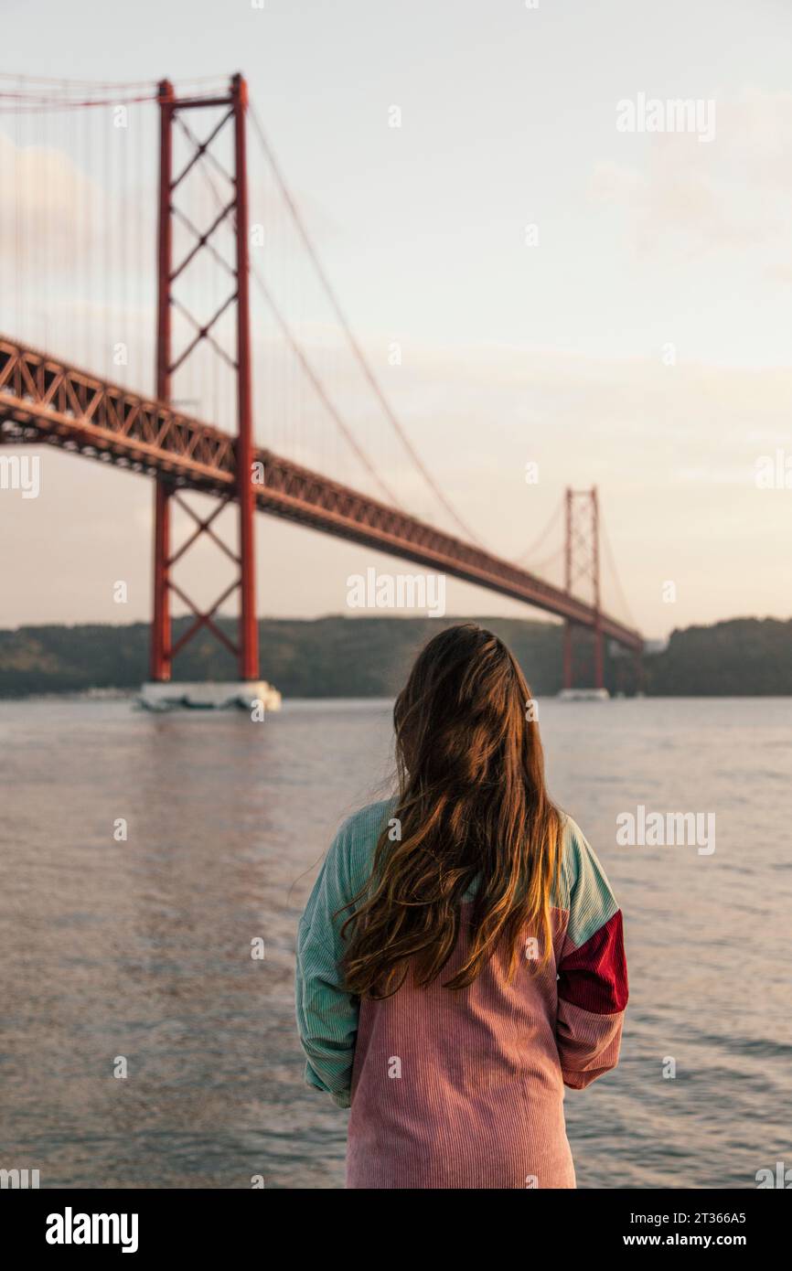 Junge Frau, die den Fluss Tejo mit der Brücke am 25. April bei Sonnenuntergang bewundert Stockfoto