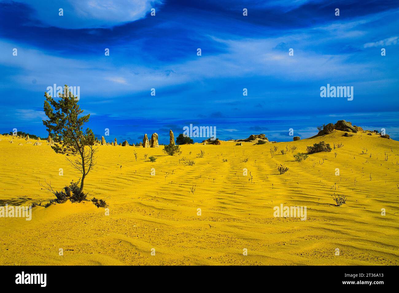 Surrealistische Wüstenlandschaft mit ockergelbem Rippensand, Felsen, Zinnen und einem harten blauen Himmel mit schimmernden Wolken Stockfoto