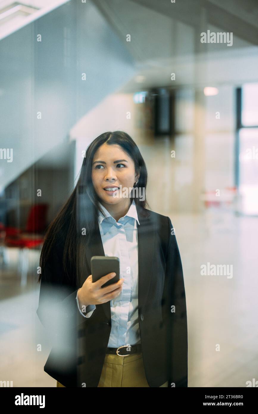 Geschäftsfrau mit Smartphone, das am Arbeitsplatz durch Glas gesehen wird Stockfoto
