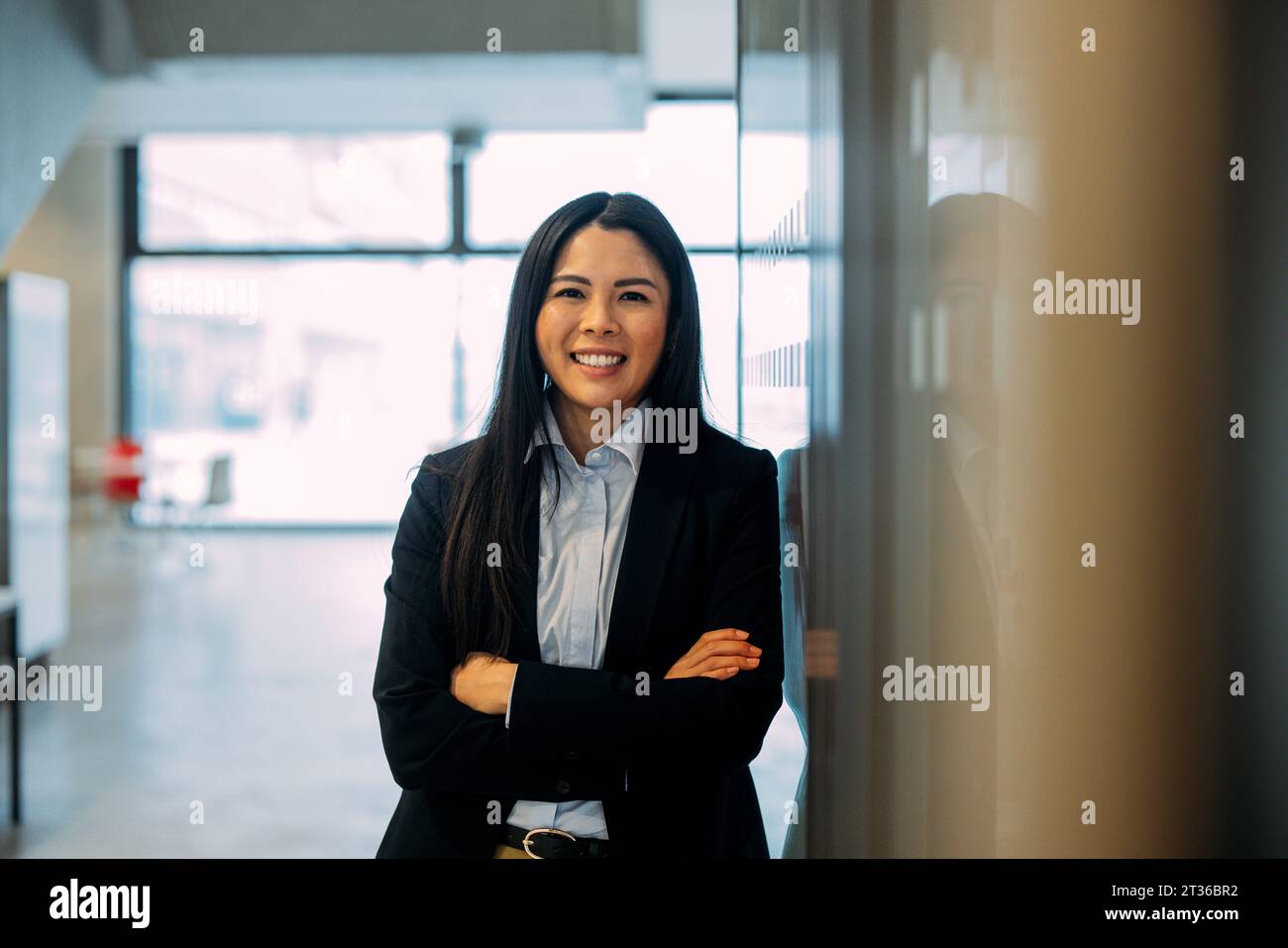Lächelnde Geschäftsfrau mit überkreuzten Armen, die am Arbeitsplatz an die Wand gelehnt ist Stockfoto