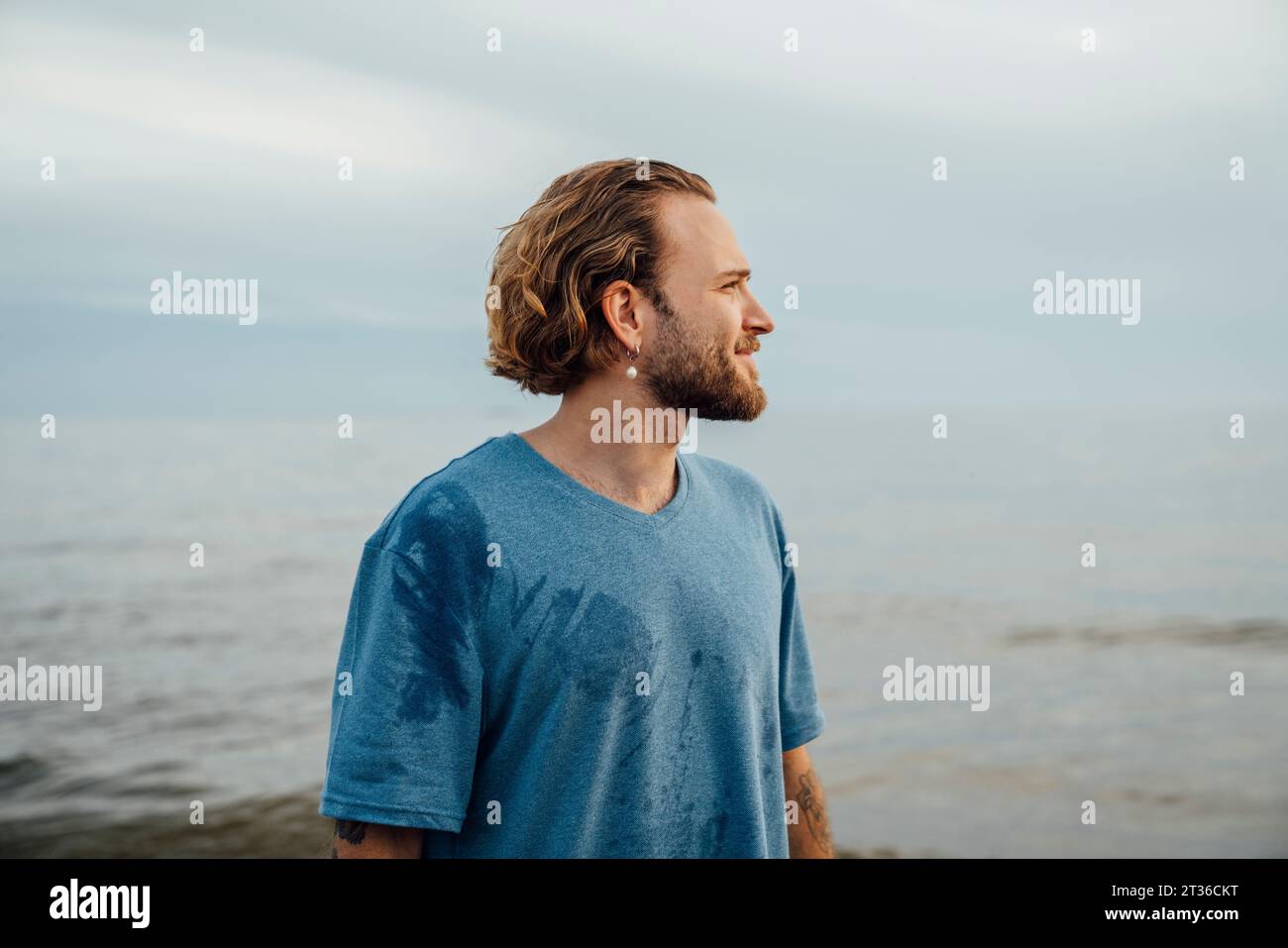 Lächelnder blonder Mann, der ein T-Shirt in der Nähe des Meeres am Strand trägt Stockfoto