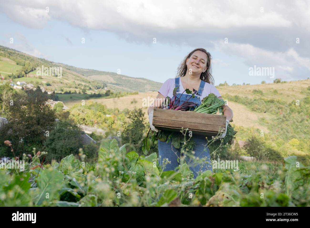 Lächelnde Frau mit Gemüsekiste unter bewölktem Himmel auf der Farm Stockfoto