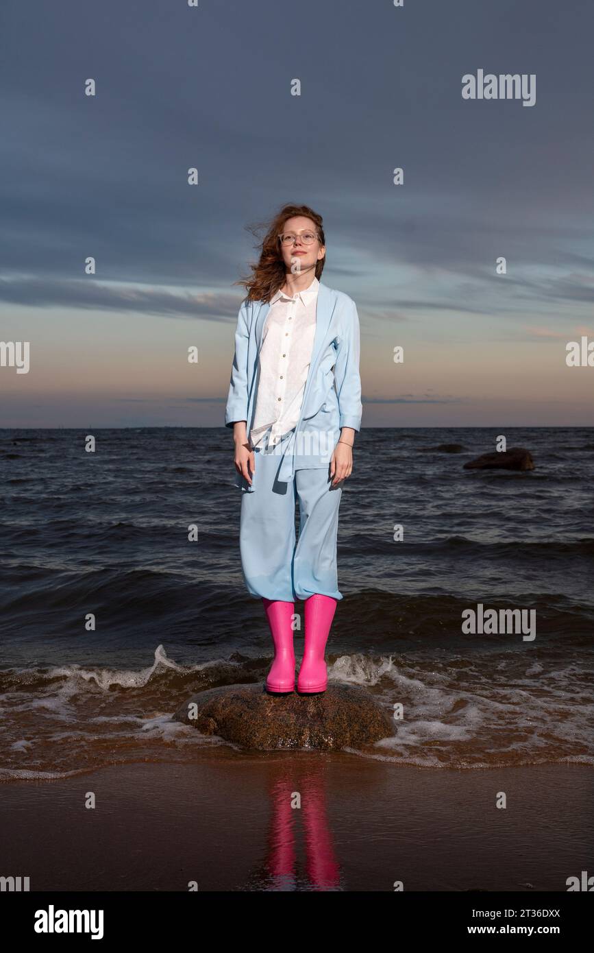 Eine junge Frau mit rosa Gummistiefeln, die bei Sonnenuntergang auf dem Felsen steht Stockfoto