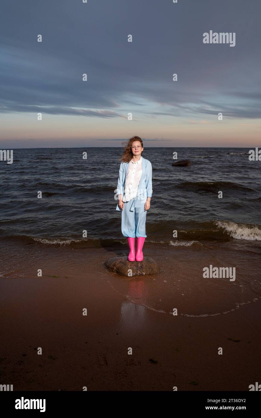 Frau mit rosafarbenen Gummistiefeln, die bei Sonnenuntergang auf dem Felsen steht Stockfoto