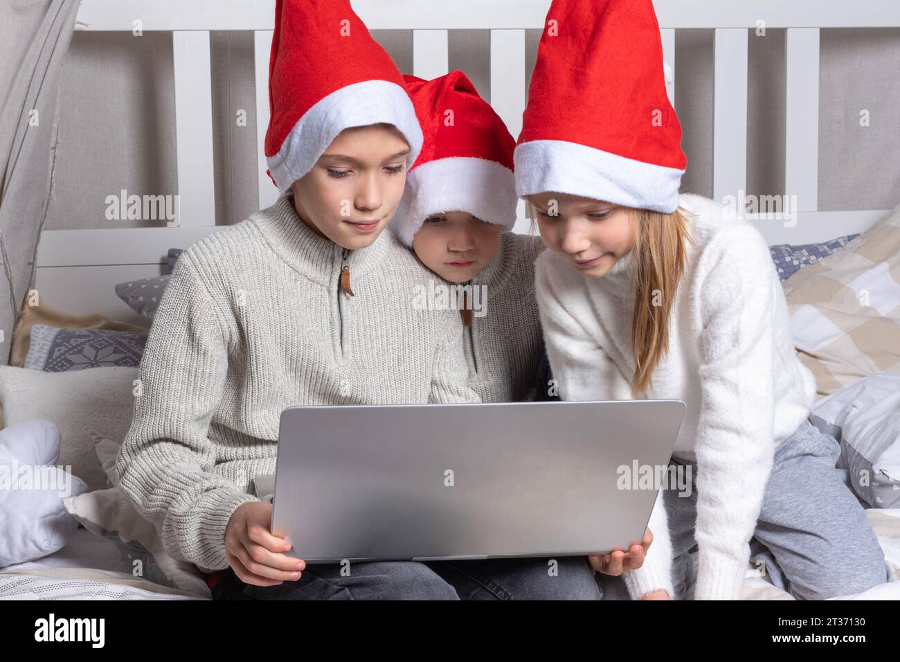 Fröhliche Kinder in roten Weihnachtsmannhüten mit einem Laptop an Heiligabend zu Hause. Jungs und ein Mädchen gratulieren online Verwandten, die auf dem Bett sitzen. Familie x-Mas ev Stockfoto