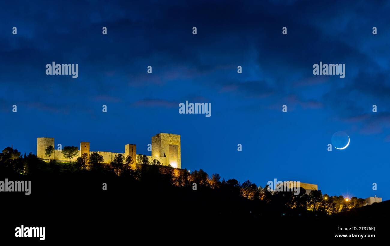 Schloss Santa Catalina in Jaen, Spanien, bei Nacht beleuchtet, mit einem bewölkten blauen Himmel und dem wachsenden Halbmond auf der rechten Seite des Bildes. Stockfoto