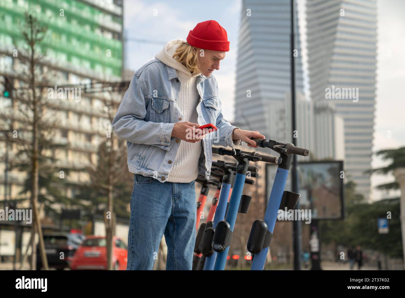 Trendiger Teenager mit mobiler App, um E-Scooter zu mieten, der auf dem Straßenparkplatz steht Stockfoto