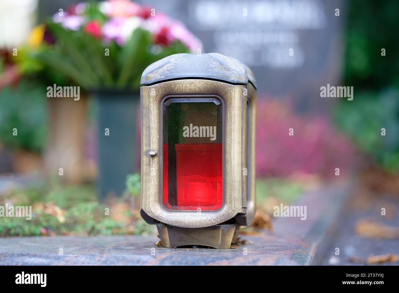 Nahaufnahme der Grablampe aus Messing mit roter Kerze und Blumenstrauß und Grabstein in verschwommenem Hintergrund Stockfoto