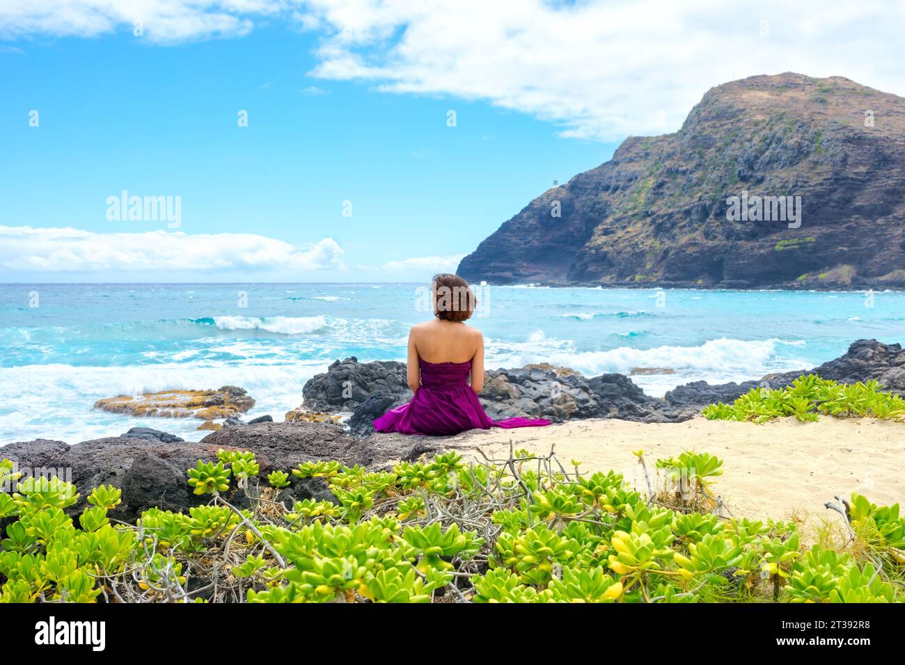 Eine junge Frau im lila Kleid sitzt am felsigen Ufer und blickt auf den Ozean am Makapu'u Beach in Oahu, Hawaii Stockfoto