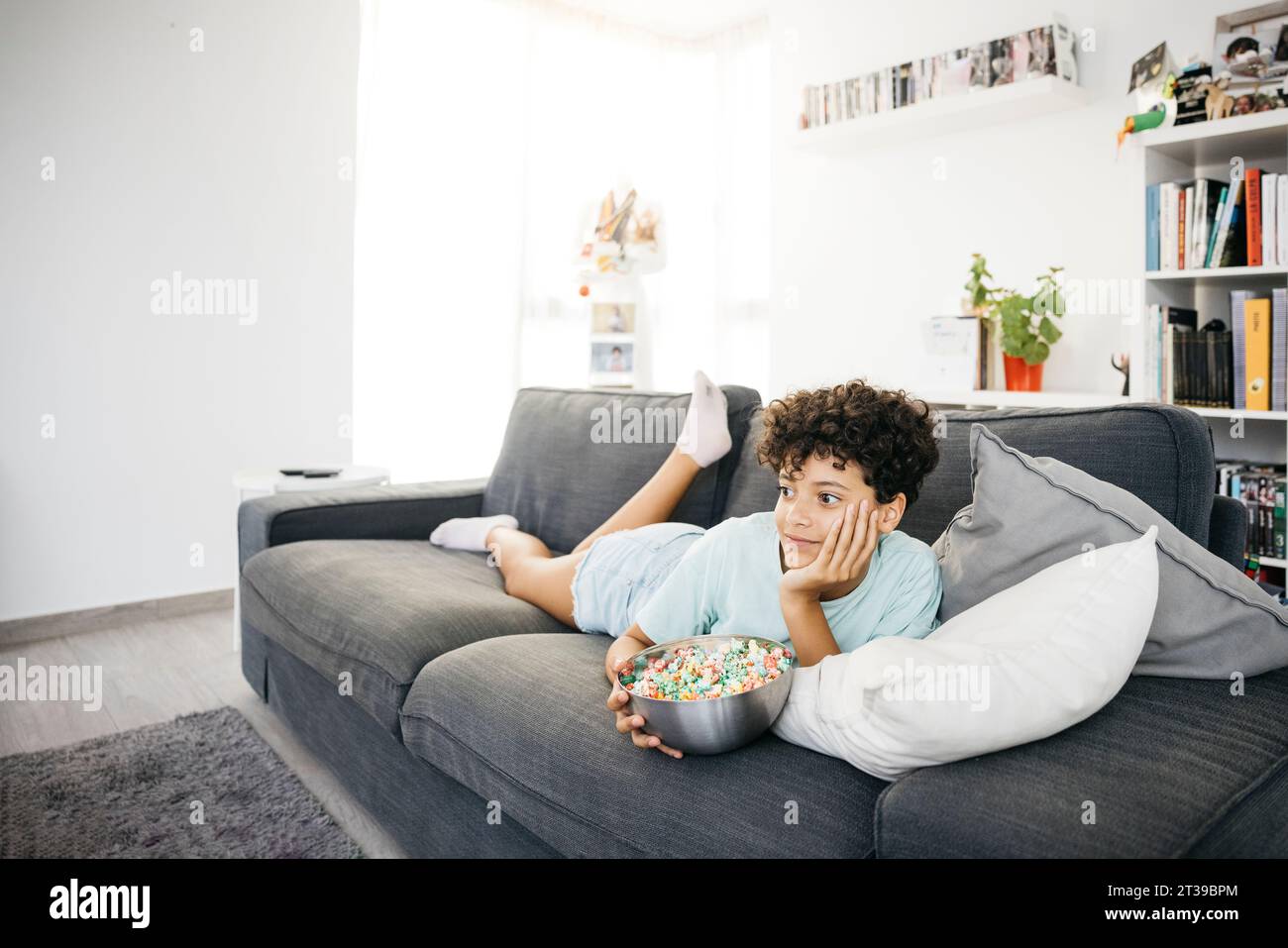 Ein zehnjähriges langweiliges Mädchen, das auf einem Sofa liegt und fernsehen sieht, mit einem tv-Controller, in einem Wohnzimmer. Ein Mädchen, das zu Hause einen Film ansieht. Stockfoto