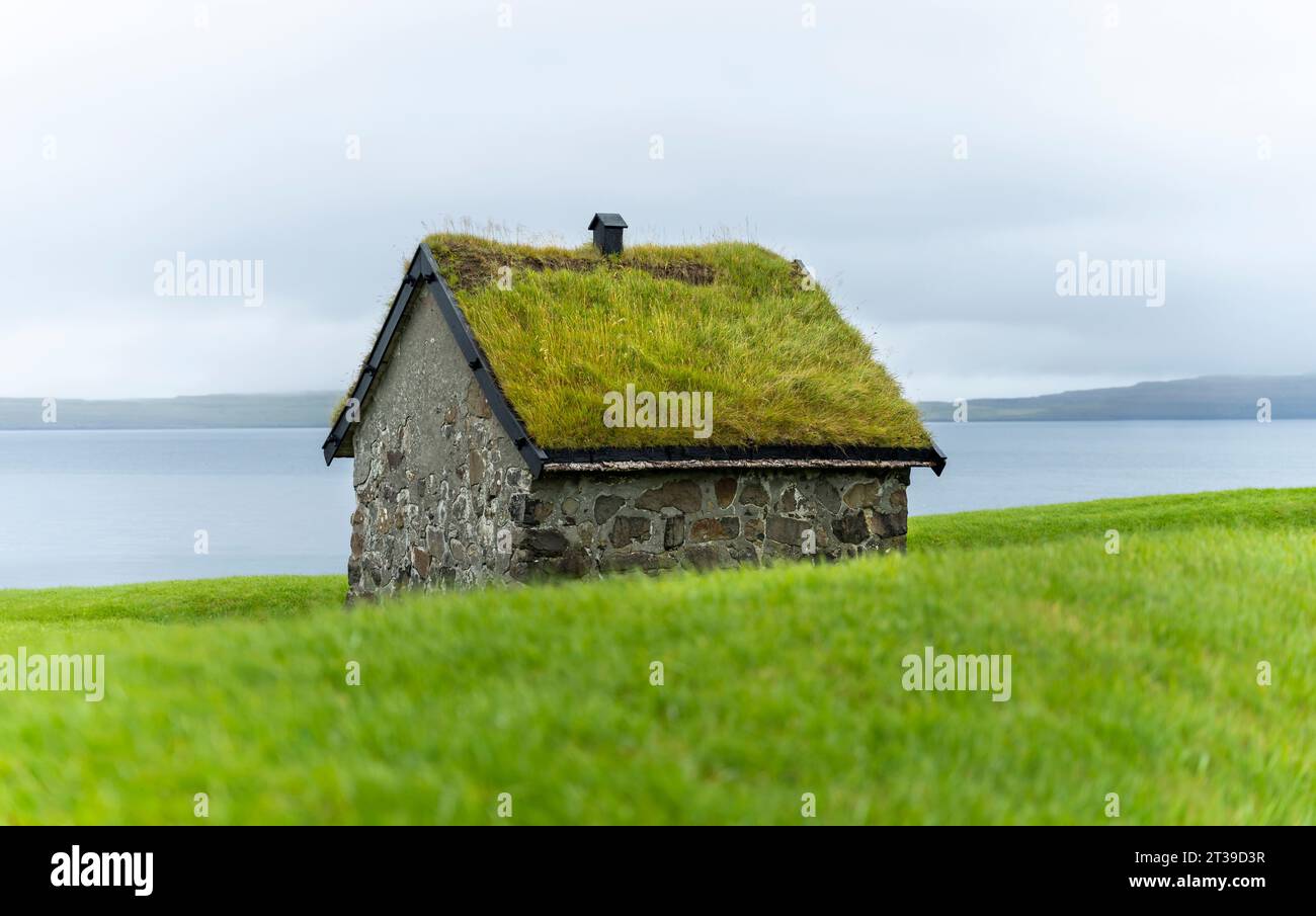 Altes Steinhaus mit grasbewachsenem Dach am grünen Ufer des ruhigen Meeres vor düsterem Himmel auf den Färöern Stockfoto