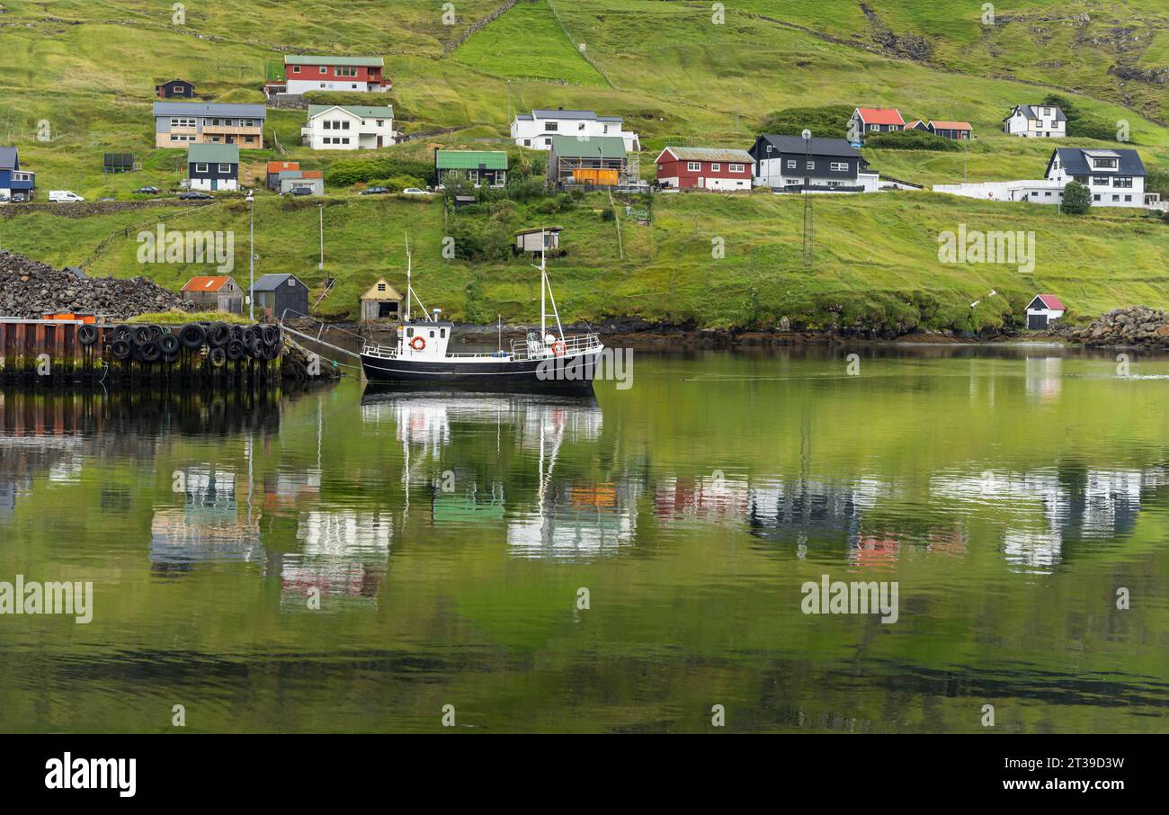 Malerischer Blick auf kleine Dorfhäuser an der grünen Küste des ruhigen Sees mit schwimmender Yacht auf Färöern Stockfoto