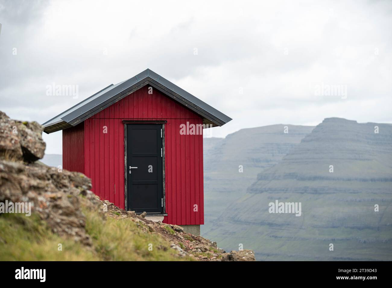 Äußere eines kleinen roten Holzhauses auf einem grasbewachsenen Hügel vor bewölktem Himmel auf den Färöer Inseln Stockfoto