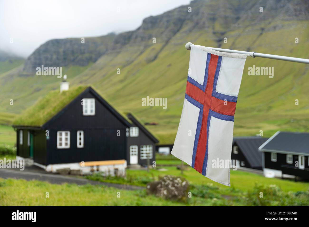 Die Nationalflagge der Färöer hängt an schwarzen Häusern, die sich auf einem Hügel in der Nähe des mit Gras bedeckten Bergrücken befinden Stockfoto