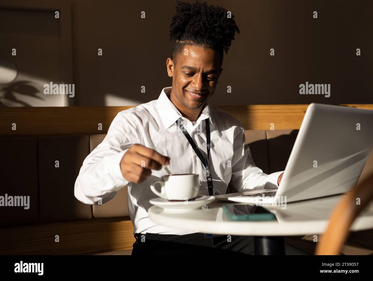 Glücklicher junger afroamerikanischer Freelancer in lässigen Klamotten und rasierten Dreadlocks, der am Kaffeetisch mit einer Tasse Kaffee sitzt, während er im Laptop surft Stockfoto