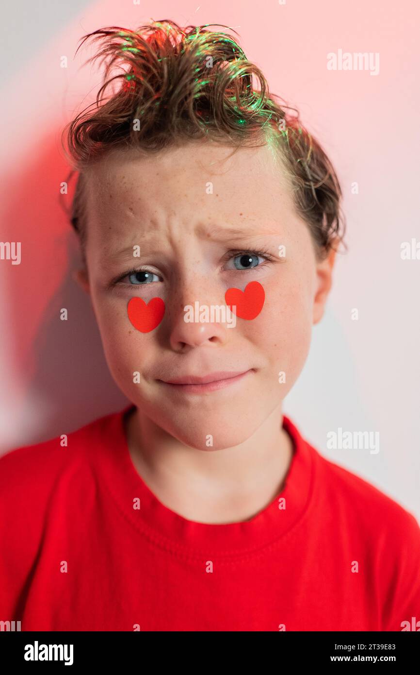 Ein kleiner Junge mit lockigen Haaren und herzförmigen Aufklebern auf den Wangen schaut ruhig in die Kamera und trägt ein rotes Hemd mit einem subtilen Hauch von abstraktem Rot Stockfoto